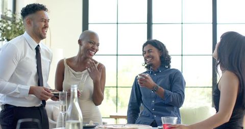 Diverse group laughing and networking in sunlit lounge with drinks and snacks