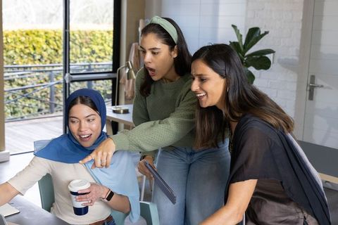Diverse female team collaborating around laptop with hijab, tablet and coffee workspace