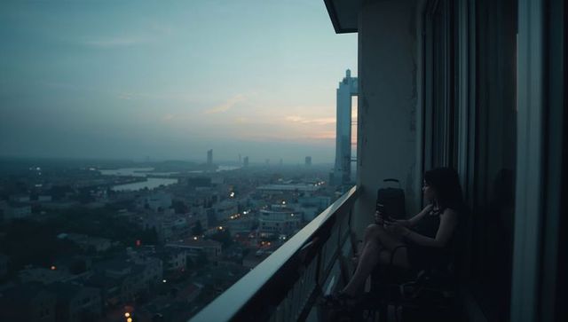 Sitting woman on high-rise balcony holding cup at dusk overlooking river city skyline lights