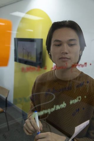 Adult asian man writing on glass board in modern workspace brainstorming and planning