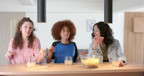 Three friends laughing and grabbing chips at modern kitchen counter during casual hangout