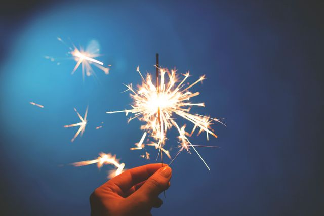 Hand holding burning sparkler against vibrant blue background. Ideal for use in promotional materials for holiday events, New Year's Eve celebrations, parties, or festive announcements.