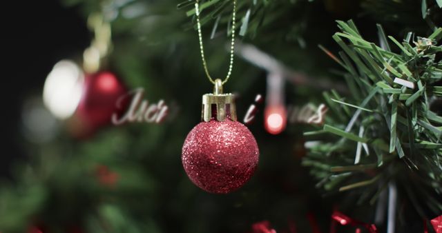 Red Christmas ornament hanging on a green tree, captured in a close-up shot. Glitter, reflecting light, encapsulates holiday spirit and enriches festive home atmosphere. Gaelic phrases in blurred background boost seasonal charm. Excellent for use in holiday greeting cards, advertisements for decoration items, or other Christmas-themed promotional materials.