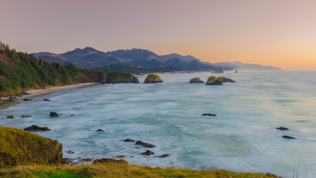 This depiction of an early morning sunrise over a coastal area features tidal waves and fog-clad mountains. Ideal for travel brochures, nature magazines, environmental posters, and websites promoting coastal tourism and outdoor activities. It conveys a serene and peaceful atmosphere, making it suited for uses related to relaxation and natural beauty.