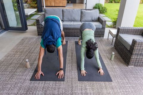 Couple practicing downward dog on covered patio with wicker sofa and matching yoga mats