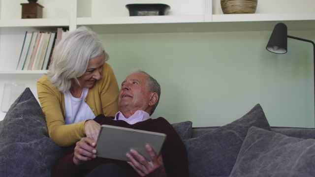 Senior couple engaged in using a tablet while relaxing on a sofa. Their focused expressions convey determination and teamwork in navigating technology. Ideal for depicting elderly connections with modern technology, warmth in senior living settings, or illustrating aging and adaptation stories.