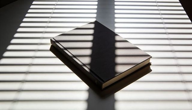 Hardcover book receiving striped sunlight through blinds on minimalist tabletop