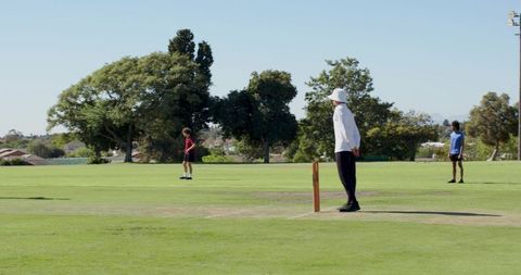 Cricket umpire standing beside stumps on sunny park outfield with distant fielders