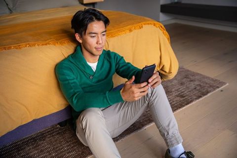 Asian man relaxing on bedroom floor leaning against bed scrolling smartphone wearing green sweater
