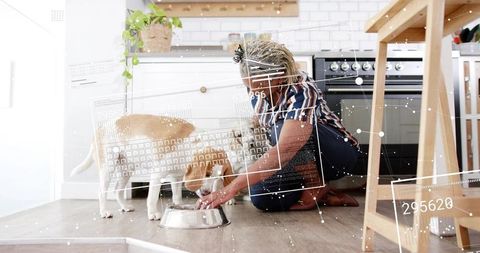 Elderly woman kneeling feeding two tan dogs from stainless bowl in bright modern kitchen