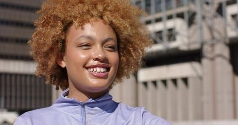 African American woman smiling in sunlit urban plaza wearing lavender zip-up jacket, casual wear