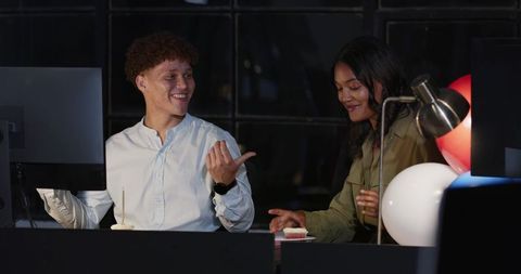 Coworkers chatting at shared desk during late-night work with cupcake and moody lighting