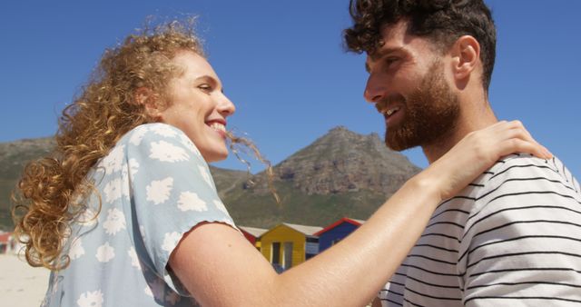 A young Caucasian couple shares a joyful moment on a sunny beach, with colorful beach huts in the background and copy space. Their smiles and affectionate embrace convey a sense of happiness and romantic connection.