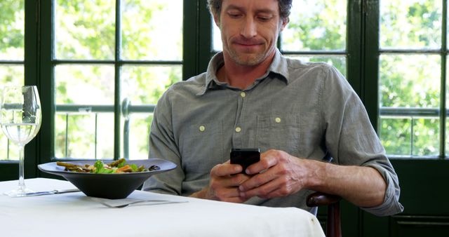 Man enjoying his meal while looking at his phone in a cozy restaurant. Ideal for scenes of digital technology use during meal times, solo dining experiences, relaxation in food establishments, casual lifestyle moments, and modern dining habits.