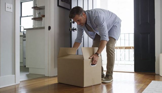 Middle-aged man unpacking cardboard box at sunlit home entryway with hardwood floors