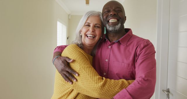 Happy senior couple standing indoors, smiling and embracing each other. This image can be ideal for depicting themes of love, companionship, interracial relationships, and positive emotional expressions. Perfect for use in marketing materials, advertisements targeting senior living and healthcare services, or articles revolving around healthy aging and relationships.