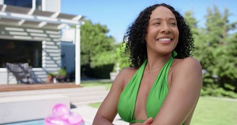 Smiling woman wearing green swimsuit standing poolside on sunny backyard deck, copy space
