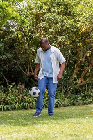African american man balancing soccer ball in lush backyard garden wearing jeans