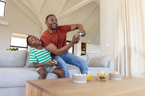 African american father and son playing video games and snacking on sofa in sunlit home