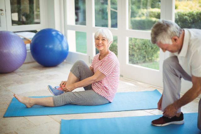 Senior couple enjoying a moment of relaxation after exercising at home. They are sitting on yoga mats, smiling and removing their shoes. Ideal for use in articles or advertisements about senior fitness, healthy aging, active lifestyles, and wellness programs for the elderly.
