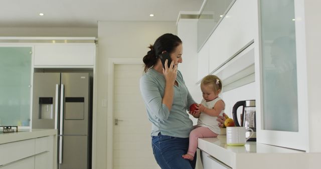 Caucasian mother holding her baby talking on smartphone in the kitchen at home. motherhood, love and childcare concept