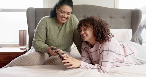 Smiling friends sharing smartphone on sunlit cozy bed wearing knit sweaters and loungewear