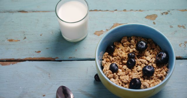 A bowl of cereal with blueberries is accompanied by a glass of milk on a rustic wooden table, with copy space. This setup suggests a healthy and simple breakfast choice.