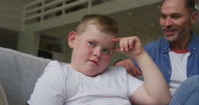 Young boy sitting on sofa looking thoughtful while his father smiles nearby. Ideal for content about family bonding, parenting advice, emotional intelligence in children, and creating happy family environments. Can also be used for health and wellness articles focusing on children's mental health or family lifestyle blogs.