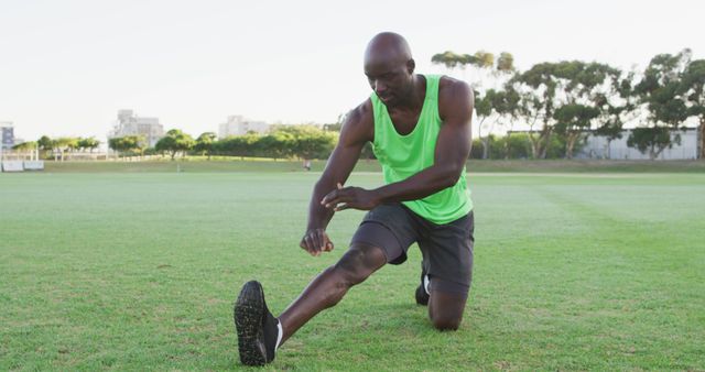 The image shows a man performing a stretching exercise on a grass field. He is wearing a bright green tank top and black shorts, indicating a sporty and active lifestyle. This image can be used for content related to fitness, aerobics, outdoor workouts, summer activities, and promoting a healthy lifestyle.