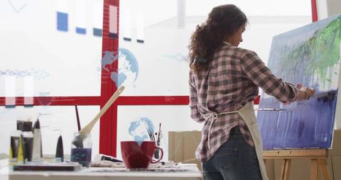 Female artist applying paint on canvas in sunlit studio with easel and palette
