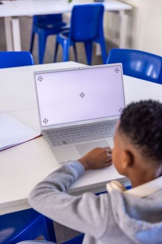 Schoolboy using laptop at classroom table with headphones and notebook for remote learning