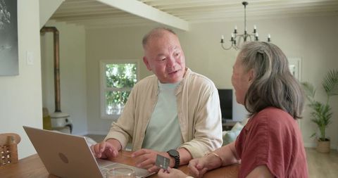 Senior asian couple managing online payment on laptop at bright rustic living room table