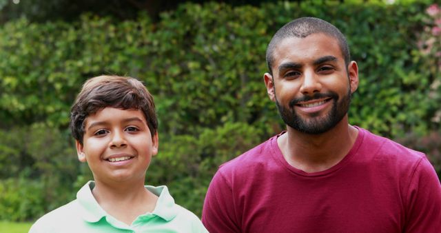 Showing a big brother and little brother spending time together in a park. They are smiling and looking towards the camera. Surrounded by green foliage, suggesting a natural and lively environment. Ideal for use in family-focused content, mental well-being discussions, or marketing involving children's interests.