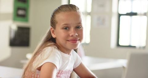 Smiling preteen girl using laptop in bright classroom with natural window backlight