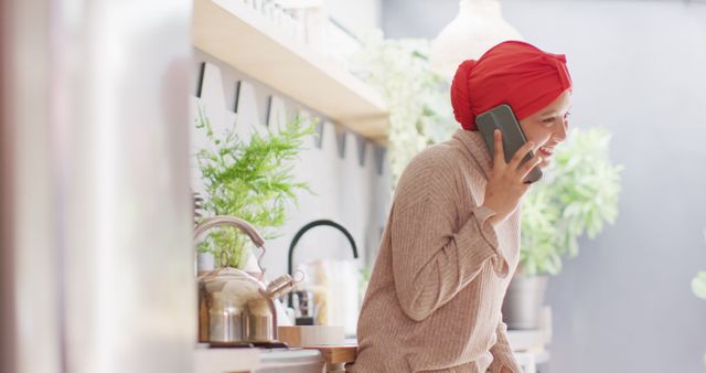 Smiling woman wearing a head scarf talking on a mobile phone in a well-lit, plant-filled kitchen. She is dressed in warm clothing and appears happy and engaged. Suitable for use in materials highlighting communication, home life, happiness, and modern lifestyle imagery.