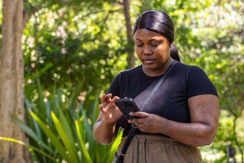 Woman checking smartphone in sunlit urban park wearing crossbody bag and olive pants