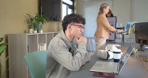 Young man working on laptop with VR headset nearby in modern open coworking office space
