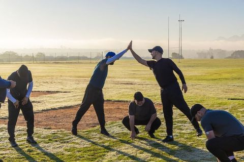 Six male teammates exchanging high-five and stretching on frosty baseball infield at dawn