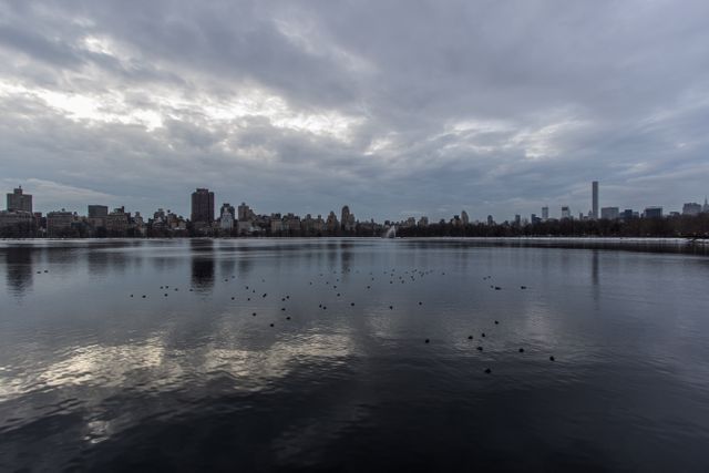 A peaceful urban skyline reflecting on a calm lake with a cloudy sky background. Ideal for use in themes related to urban tranquility, city landscapes, and nature within cities. Perfect for illustrating articles about city life, urban planning, or environmental harmony.