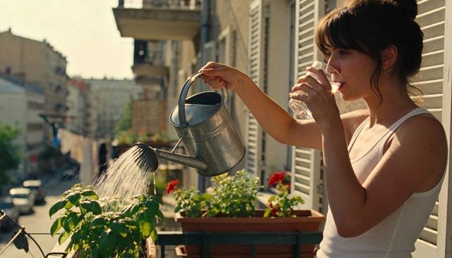 Woman watering balcony herbs while drinking water on sunlit urban apartment terrace