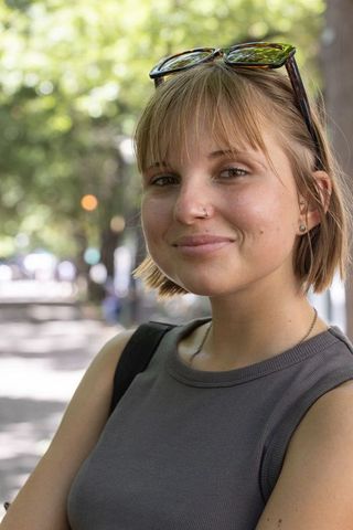 Young woman smiling on sunlit city sidewalk wearing sunglasses and nose hoop