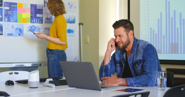 Modern office showing team working together on project. Man in foreground using laptop on call while woman in background analyzing charts on whiteboard. Ideal for depicting teamwork, business strategies, data analysis.