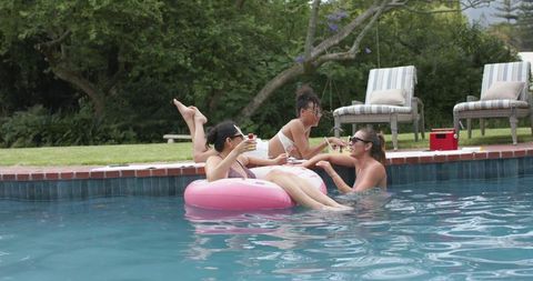 Diverse friends lounging on pink float sipping cocktails at backyard pool party