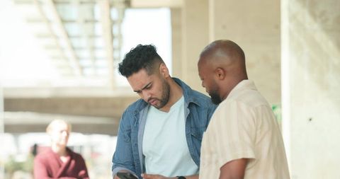 Two men checking smartphone under urban walkway, casual denim jacket and striped shirt