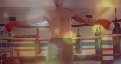 Shirtless boxer leaning on ropes, resting between rounds in moody gym with red gloves