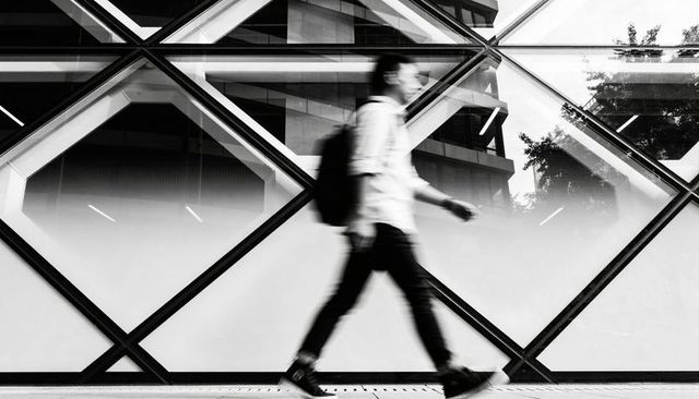 Blurry commuter walking past diamond glass facade reflecting cityscape, monochrome urban motion