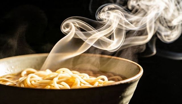 Steaming udon noodles in ceramic bowl with curling steam plumes and dark background