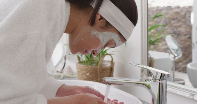 Woman with headband and bathrobe leaning over sink, practicing skincare routine. Great for articles, blogs, or websites related to beauty tips, personal care, wellness routines, and healthy skincare practices.