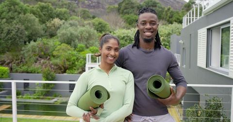 Smiling diverse couple holding green yoga mats on balcony, enjoying outdoor fitness routine