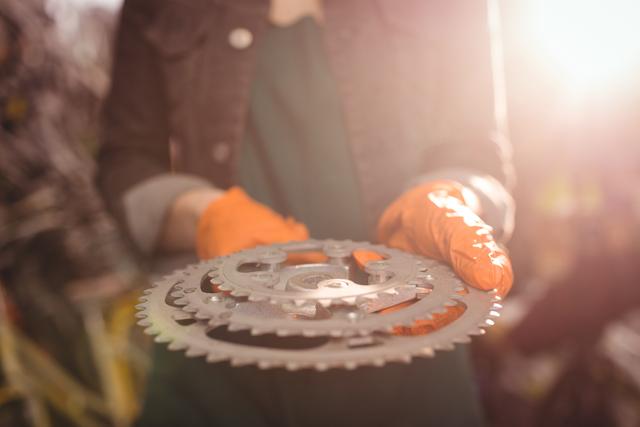 Mechanic holding bicycle gear in workshop, wearing gloves, with sunlight in background. Ideal for use in articles or advertisements related to bicycle repair, maintenance services, cycling equipment, or mechanical engineering.
